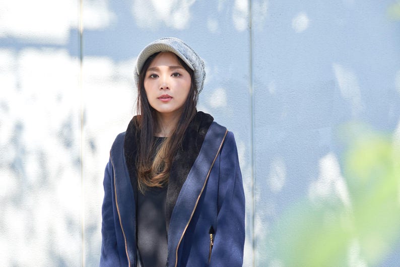 May include: A woman wearing a blue jacket with a gray knitted hat. The jacket has a zipper and a hood. The woman is standing in front of a light blue wall.