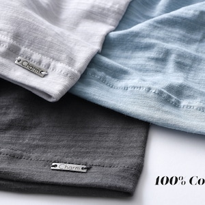 Pode incluir: Tr&ecirc;s camisetas em cores diferentes, cinza, azul e cinza escuro, com uma etiqueta prateada que diz "Charm" em cada uma. O texto "100% Algod&atilde;o" &eacute; impresso em um fundo branco.