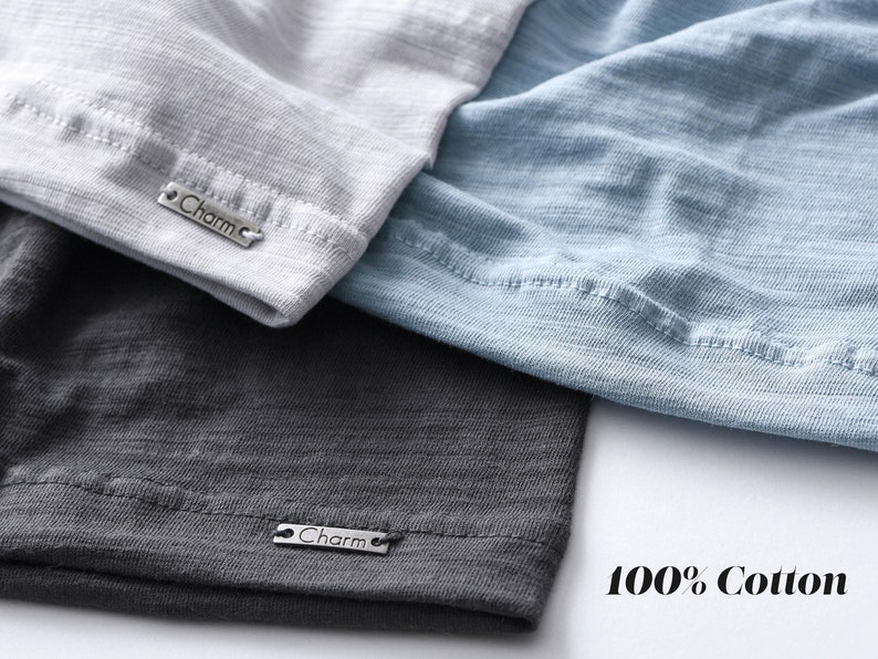 Pode incluir: Tr&ecirc;s camisetas em cores diferentes, cinza, azul e cinza escuro, com uma etiqueta prateada que diz "Charm" em cada uma. O texto "100% Algod&atilde;o" &eacute; impresso em um fundo branco.