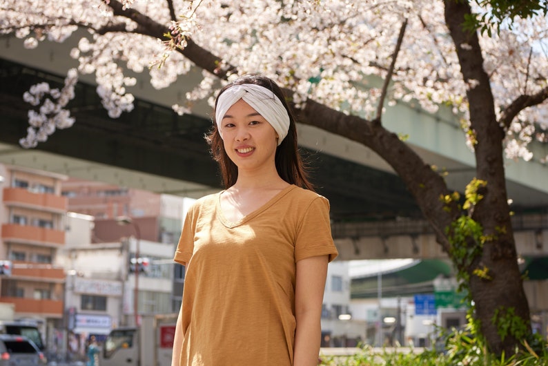 May include: A white twisted headband made of fabric worn on the head, paired with a plain mustard yellow short-sleeved v-neck t-shirt. The headband features a central knot detail and a soft, ribbed texture, providing a simple accessory.