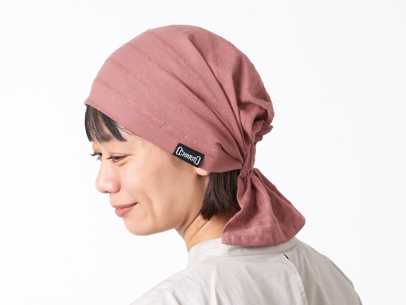 K&ouml;nnte beinhalten: Ein staubiges rosafarbenes Baumwollbandana mit einem schwarzen "CHARM'D"-Logo. Das Bandana ist am Hinterkopf zu einem Knoten gebunden.