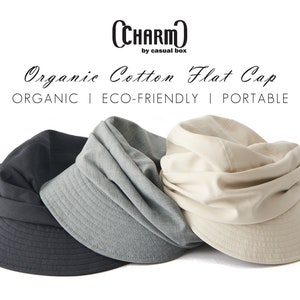 May include: Three organic cotton flat caps in charcoal, grey, and beige. The caps have a structured brim and a soft, draped crown. Text on the image reads "CHARM by casual box" and "Organic Cotton Flat Cap". The image also includes the words "ORGANIC | ECO-FRIENDLY | PORTABLE".