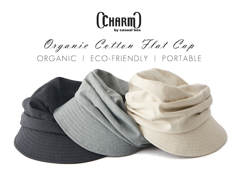 May include: Three organic cotton flat caps in charcoal, grey, and beige. The caps have a structured brim and a soft, draped crown. Text on the image reads "CHARM by casual box" and "Organic Cotton Flat Cap". The image also includes the words "ORGANIC | ECO-FRIENDLY | PORTABLE".