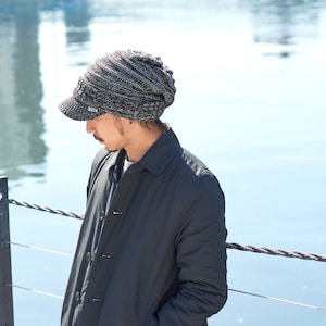 May include: A person wearing a gray and black knit beanie with a brim and a black jacket. The person is standing near a body of water.