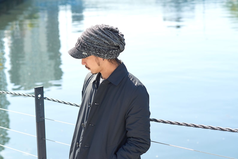 May include: A person wearing a gray and black knit beanie with a brim and a black jacket. The person is standing near a body of water.
