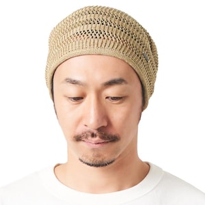 May include: A beige knit beanie featuring a textured, open-weave pattern with a solid ribbed band at the base. The hat has a rounded crown and includes a small, rectangular metallic brand tag attached to the side of the lower edge.
