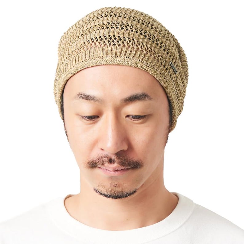 May include: A beige knit beanie featuring a textured, open-weave pattern with a solid ribbed band at the base. The hat has a rounded crown and includes a small, rectangular metallic brand tag attached to the side of the lower edge.