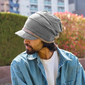 May include: A gray, soft fabric hat with a brim. The hat has a layered design and a brown band around the brim. The person in the image is wearing a white t-shirt and a denim jacket.
