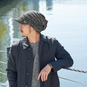 May include: A charcoal gray and black marled knit newsboy cap featuring a short, stiff brim and a slouchy, textured crown. The hat is constructed from a thick, ribbed yarn material and includes a small, rectangular white brand tag on the brim.