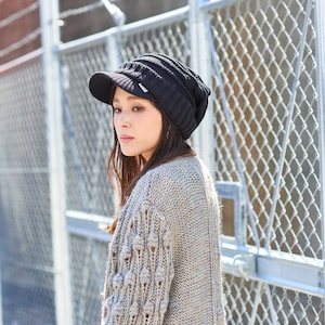 May include: A woman wearing a black knit beanie with a visor and a beige knitted cardigan with textured details. She is standing in front of a chain link fence.