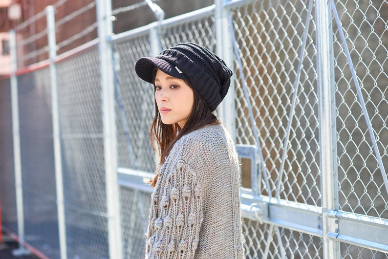 May include: A woman wearing a black knit beanie with a visor and a beige knitted cardigan with textured details. She is standing in front of a chain link fence.