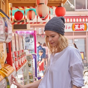 May include: A dark gray knit beanie with a textured, ribbed pattern worn on the head. The beanie features a close-fitting design with a slightly slouchy silhouette, crafted from a fine yarn material that displays a consistent, repeating stitch detail.