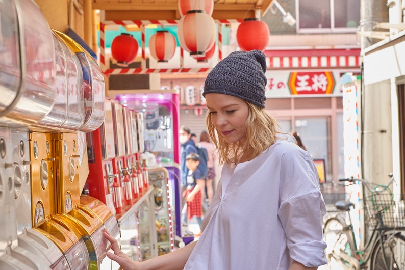 May include: A dark gray knit beanie with a textured, ribbed pattern worn on the head. The beanie features a close-fitting design with a slightly slouchy silhouette, crafted from a fine yarn material that displays a consistent, repeating stitch detail.