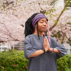 May include: A woman with long black braids and a purple headband is meditating with her hands clasped in front of her. She is wearing a gray shirt and has pink and purple beads on her wrist. The background is a blurred image of pink cherry blossoms.