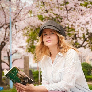 K&ouml;nnte beinhalten: Eine Frau in einem wei&szlig;en Hemd und einer dunkelgrauen Flatcap sitzt unter einem Kirschbl&uuml;tenbaum. Sie h&auml;lt ein Taschenbuch mit dem Titel "Sharp Objects" von Gillian Flynn.