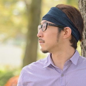 May include: A man wearing a dark blue headband and black framed glasses, looking off to the side. He is wearing a light purple button-down shirt.