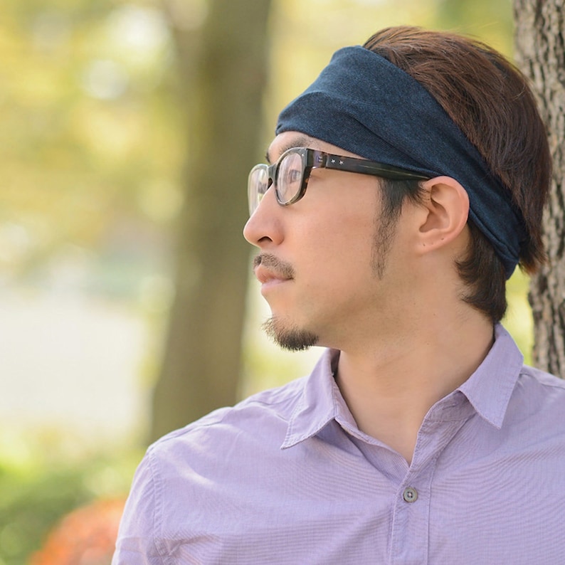 May include: A man wearing a dark blue headband and black framed glasses, looking off to the side. He is wearing a light purple button-down shirt.