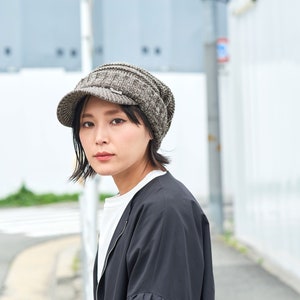 May include: A woman wearing a gray and black knit newsboy cap with a visor. She is wearing a black jacket and a white shirt.