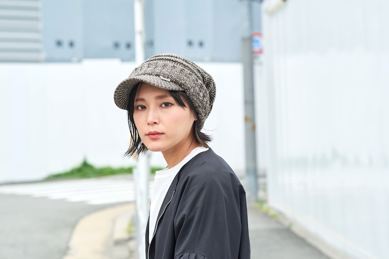May include: A woman wearing a gray and black knit newsboy cap with a visor. She is wearing a black jacket and a white shirt.