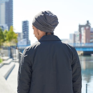 Pode incluir: Uma pessoa usando um gorro cinza e uma jaqueta preta est&aacute; em p&eacute; em um ambiente urbano. A pessoa est&aacute; de costas para a c&acirc;mera e olhando para um corpo d'&aacute;gua.