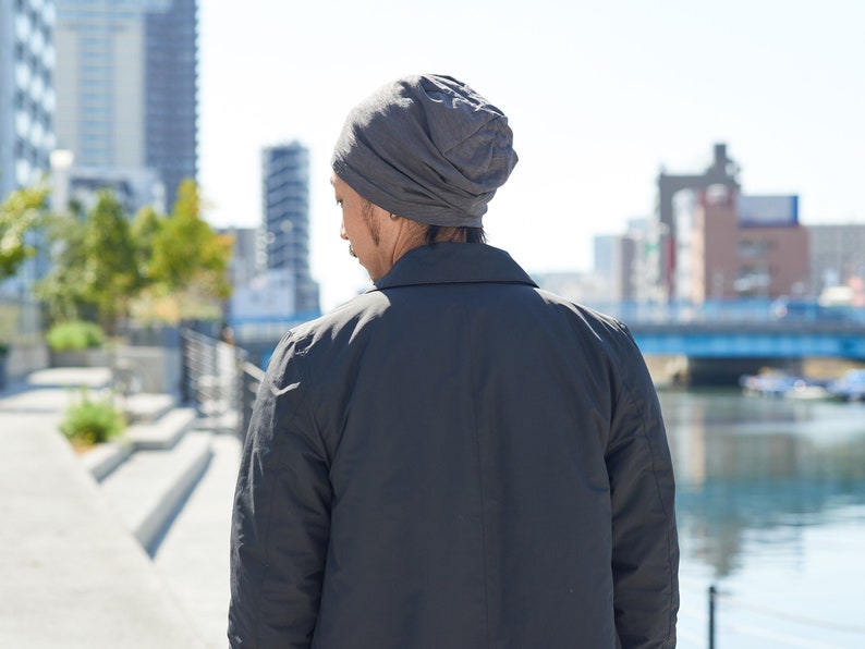 Pode incluir: Uma pessoa usando um gorro cinza e uma jaqueta preta est&aacute; em p&eacute; em um ambiente urbano. A pessoa est&aacute; de costas para a c&acirc;mera e olhando para um corpo d'&aacute;gua.