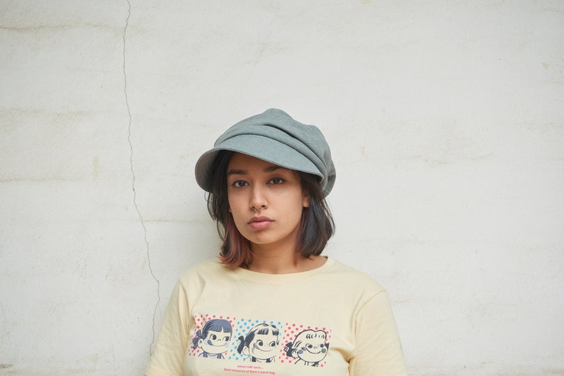 May include: A pale grey newsboy cap with a rounded crown and a small peak. The person is wearing a light yellow t-shirt with a cartoon graphic of three girls. The background is a white wall with a vertical crack.
