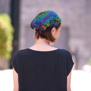 Puede incluir: Un pa&ntilde;uelo colorido con un patr&oacute;n vibrante de color turquesa, morado y amarillo. La persona que lo lleva viste una camiseta negra. El fondo est&aacute; borroso, lo que sugiere un entorno al aire libre.