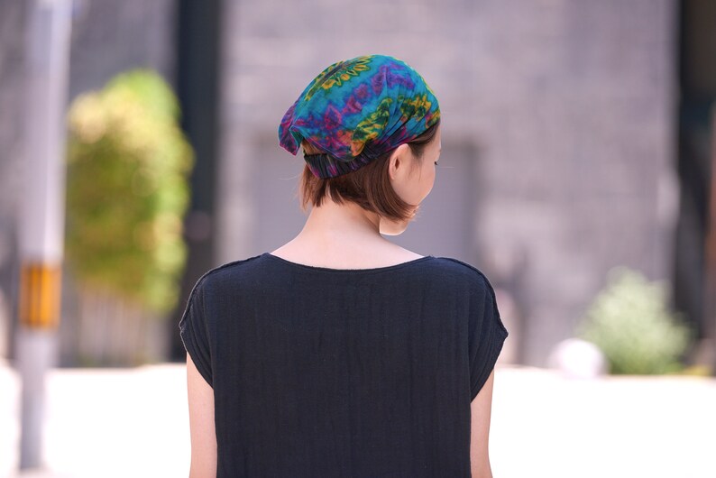 Puede incluir: Un pa&ntilde;uelo colorido con un patr&oacute;n vibrante de color turquesa, morado y amarillo. La persona que lo lleva viste una camiseta negra. El fondo est&aacute; borroso, lo que sugiere un entorno al aire libre.