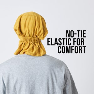 K&ouml;nnte beinhalten: Ein gelber Stoffhut mit einem Gummiband am unteren Rand. Der Hut wird auf dem Kopf einer Person getragen. Der Text "NO-TIE ELASTIC FOR COMFORT" ist auf wei&szlig;em Hintergrund gedruckt.
