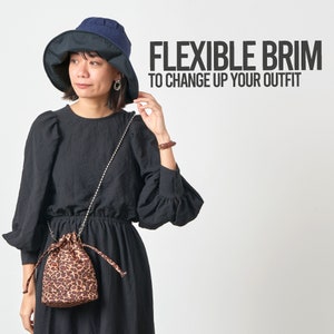 May include: A woman wearing a black dress and a blue and black hat with a flexible brim. The hat has a wide brim that can be folded up or down. The woman is also carrying a small leopard print bag with a silver chain strap. The text "FLEXIBLE BRIM TO CHANGE UP YOUR OUTFIT" is displayed above the woman.