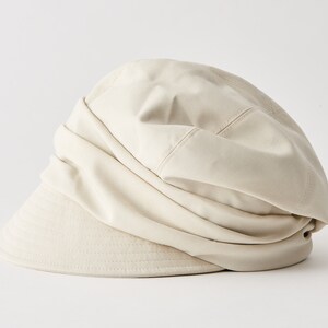 May include: A beige fabric hat with a rounded crown and a short, structured brim. The hat's design features a gathered, draped effect on the crown, creating a soft, textured appearance. The hat is a neutral colour, suitable for various outfits.