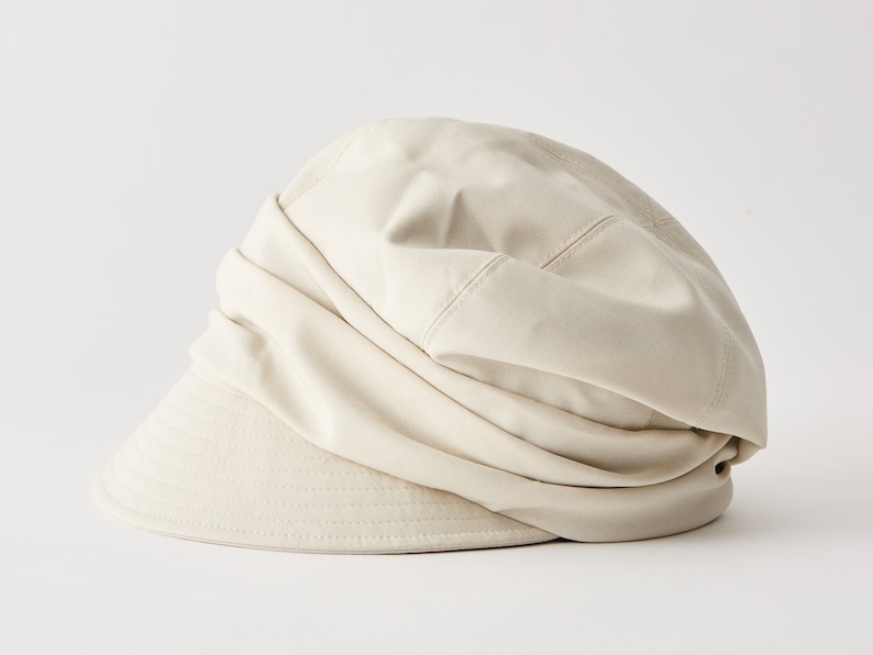 May include: A beige fabric hat with a rounded crown and a short, structured brim. The hat's design features a gathered, draped effect on the crown, creating a soft, textured appearance. The hat is a neutral colour, suitable for various outfits.