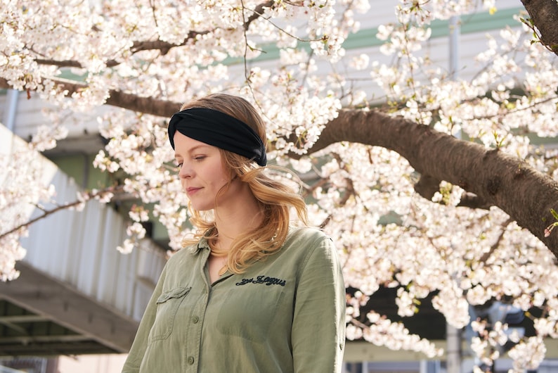 May include: A solid black fabric headband featuring a twisted knot detail at the center front. The headband is worn around the head, resting against the hair, and is constructed from a smooth, flexible material that conforms to the shape of the head.