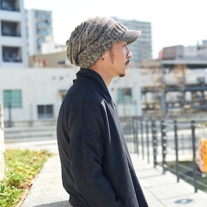May include: A man wearing a brown and gray knitted beanie with a brim, standing in front of a stone wall and looking to the right.