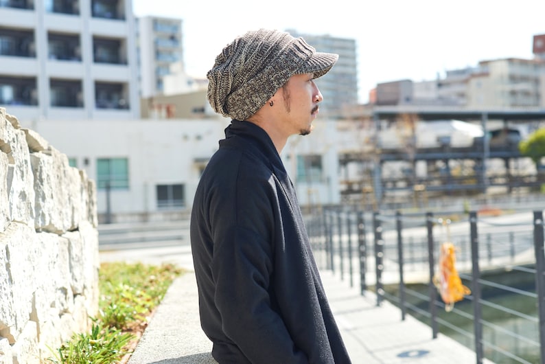 May include: A man wearing a brown and gray knitted beanie with a brim, standing in front of a stone wall and looking to the right.