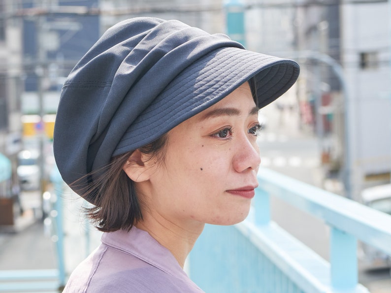 May include: A dark grey hat with a wide brim and a gathered crown. The hat is made of a soft, possibly fabric material. The person in the image is wearing a light purple shirt.