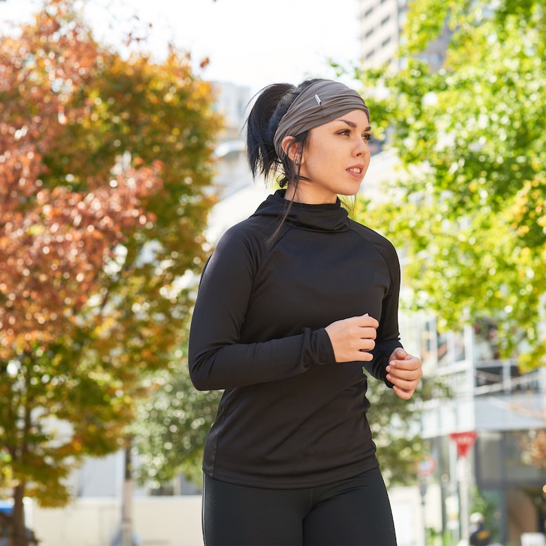 May include: A woman wearing a black mock neck shirt and a gray headband is jogging outdoors. She is wearing black leggings and has a determined expression on her face.