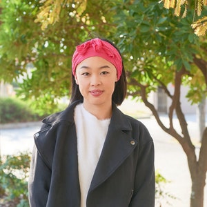 May include: A woman with short black hair is wearing a black jacket, a white sweater, and a pink headband. She is standing in front of a tree with green leaves.
