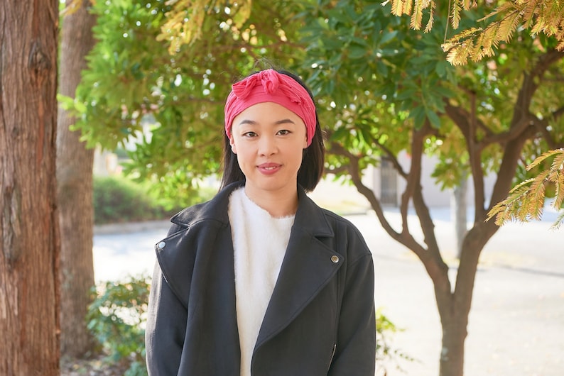 May include: A woman with short black hair is wearing a black jacket, a white sweater, and a pink headband. She is standing in front of a tree with green leaves.