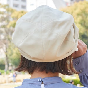 May include: A cream-coloured beret made of a leather-like material, featuring stitched detailing. The hat is being adjusted by someone wearing a blue long-sleeved shirt. The background is a blurred outdoor scene.