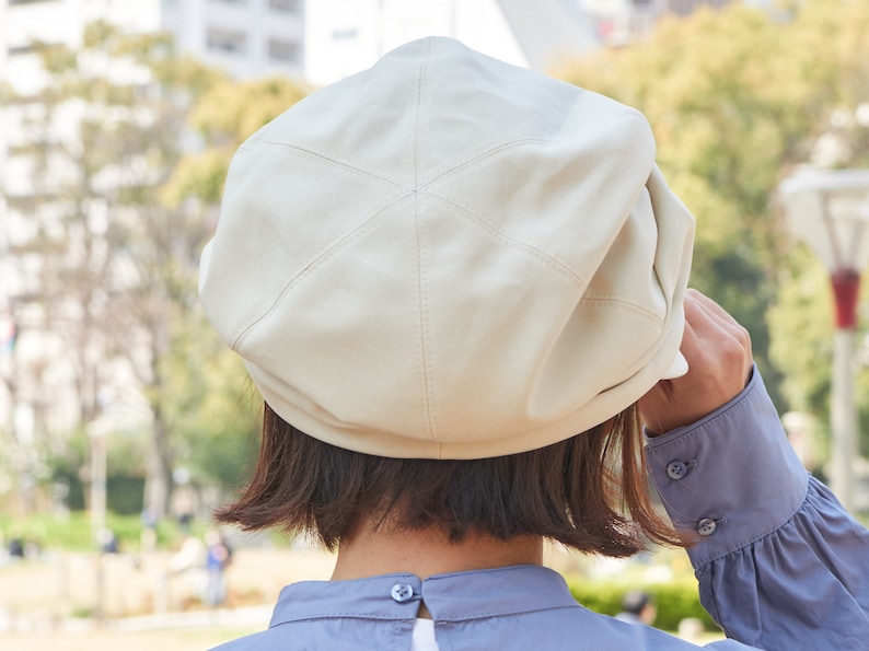 May include: A cream-coloured beret made of a leather-like material, featuring stitched detailing. The hat is being adjusted by someone wearing a blue long-sleeved shirt. The background is a blurred outdoor scene.