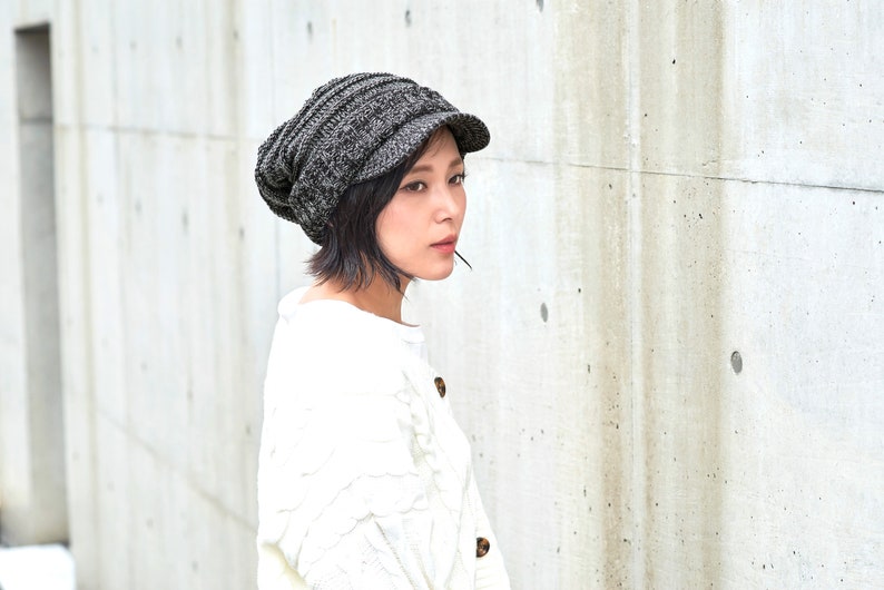 May include: A woman wearing a gray knitted beanie with a brim and a white sweater.