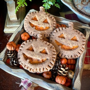 Puede incluir: Tres galletas con forma de calabaza con caras de jack-o'-lantern talladas se encuentran en una bandeja de madera llena de pequeñas calabazas y piñas. Las galletas son de color marrón claro con relleno naranja.