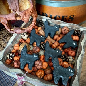 Puede incluir: Una bandeja de madera llena de calabazas en miniatura, piñas y figuras de gatos negros con ojos blancos. Una calabaza tallada y un recipiente de madera están en el fondo. La escena evoca un tema rústico, otoñal y de Halloween.