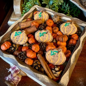 Puede incluir: Una bandeja de madera llena de decoraciones de otoño, incluyendo pequeñas calabazas naranjas, palitos de canela y piñas marrones. También hay varias galletas pequeñas con forma de calabaza con tallos verdes y glaseado marrón.