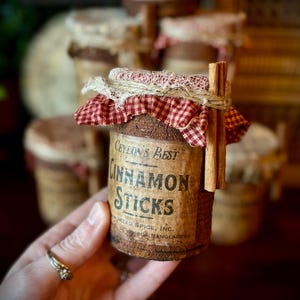 Może przedstawiać: Słoik cynamonu Ceylon's Best Cinnamon Sticks z etykietą w stylu vintage. Etykieta głosi "Cinnamon Sticks". Słoik jest owinięty czerwoną i białą tkaniną w kratkę i związany sznurkiem. W tle widoczne są inne podobne słoiki.