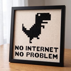 May include: A framed cross-stitch piece featuring a black pixelated dinosaur silhouette on a beige fabric background. Below the dinosaur, the text reads NO INTERNET NO PROBLEM in black, blocky, pixel-style lettering within a simple black frame.