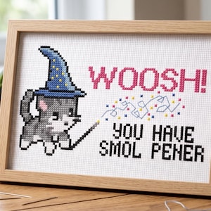 May include: A framed cross-stitch embroidery featuring a grey and white cat wearing a blue wizard hat with yellow stars. The cat holds a wand emitting blue, pink, and yellow sparkles. The text reads WOOSH! YOU HAVE SMOL PENER in pink and black.