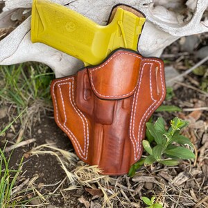 May include: A brown leather holster with white stitching, designed to hold a yellow handgun. The holster is sitting on a bed of brown dirt and green grass.