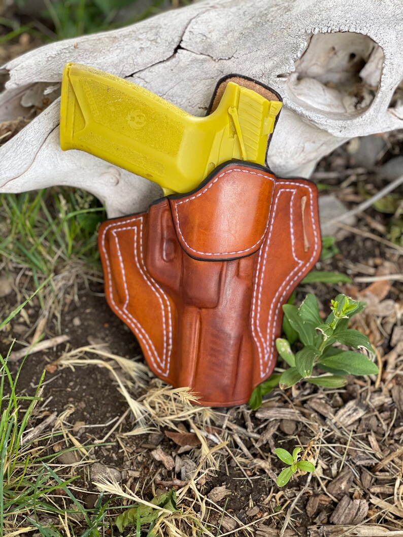 May include: A brown leather holster with white stitching, designed to hold a yellow handgun. The holster is sitting on a bed of brown dirt and green grass.